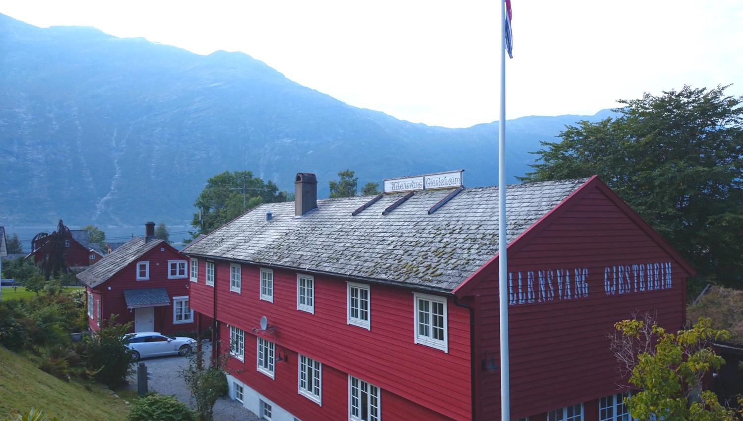 Ullensvang Gjesteheim viewed from the courtyard, featuring its red wooden building, Norwegian flag and mountain backdrop.