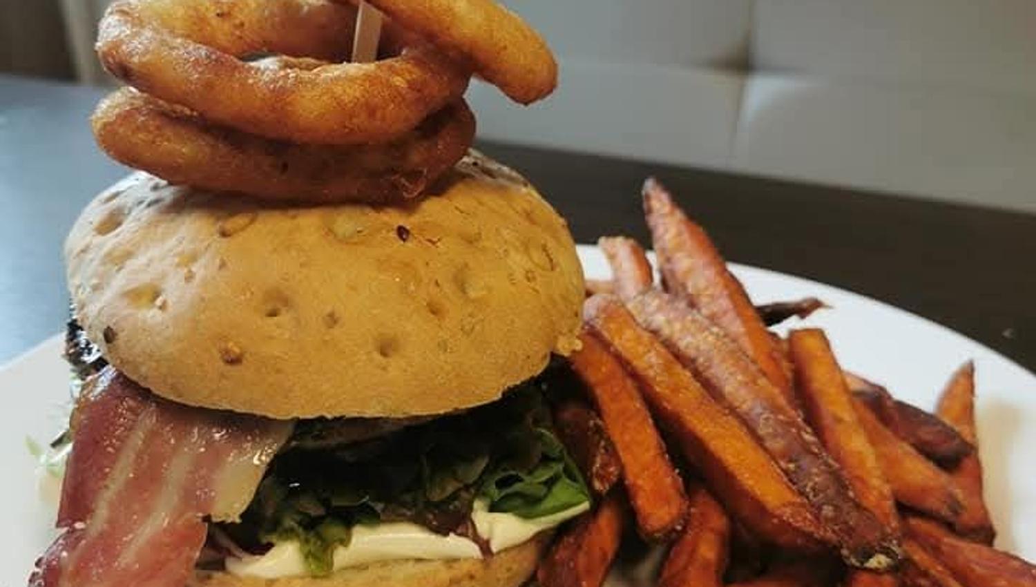 Sandwich burger topped with onion rings on a pick, served on a white plate with fries on the side.
