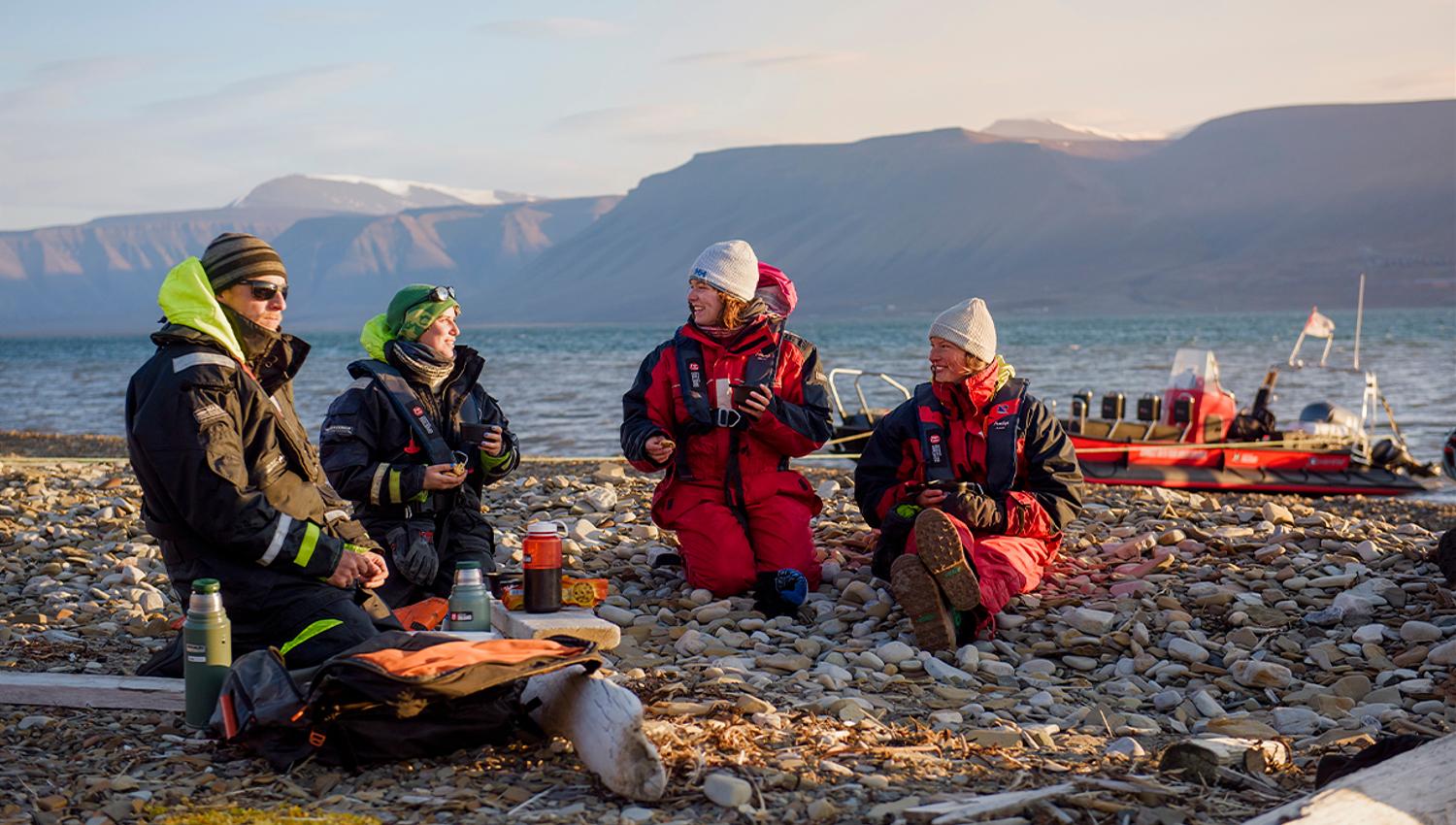 To guider og to gjester som sitter på en strand og prater med en RIB båt ankret langs sjøkanten i fjorden i bakgrunnen