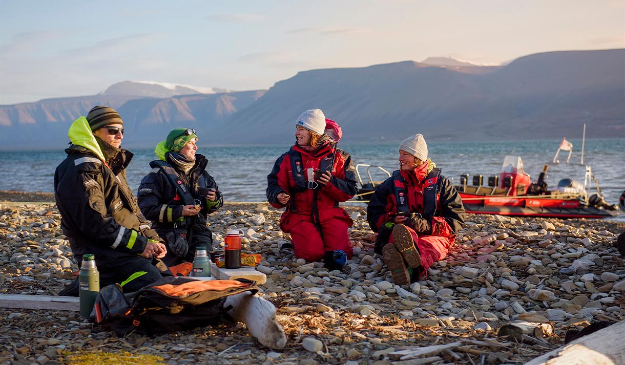 Two guides and two guests sitting on a beach talking with each other, and a RIB boat anchored by the shore of the fjord in the background