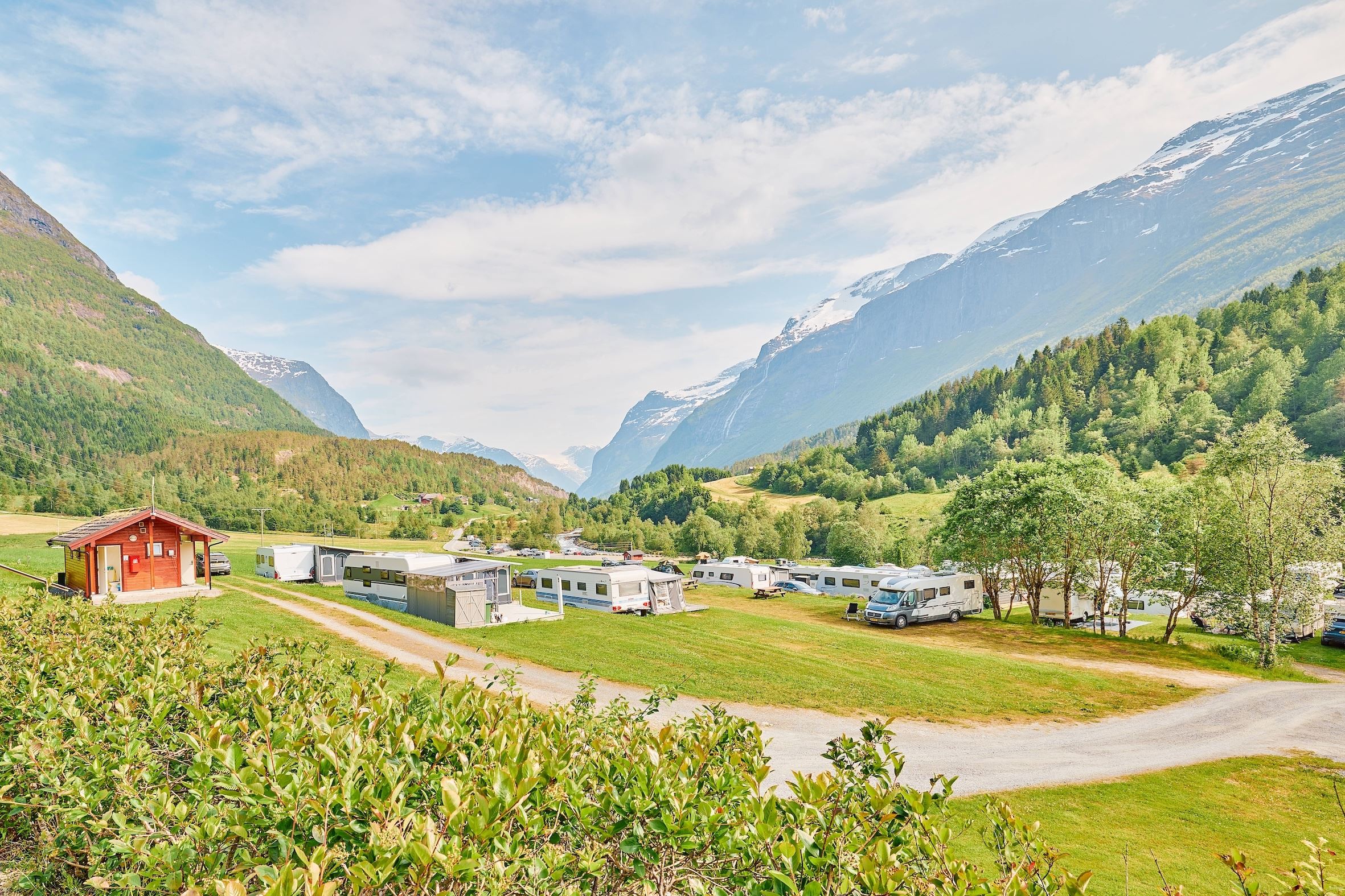 Campingplass med bubilar og campingvogner på ei grøn eng i fjelldal, med skog, høge fjell og blå himmel..