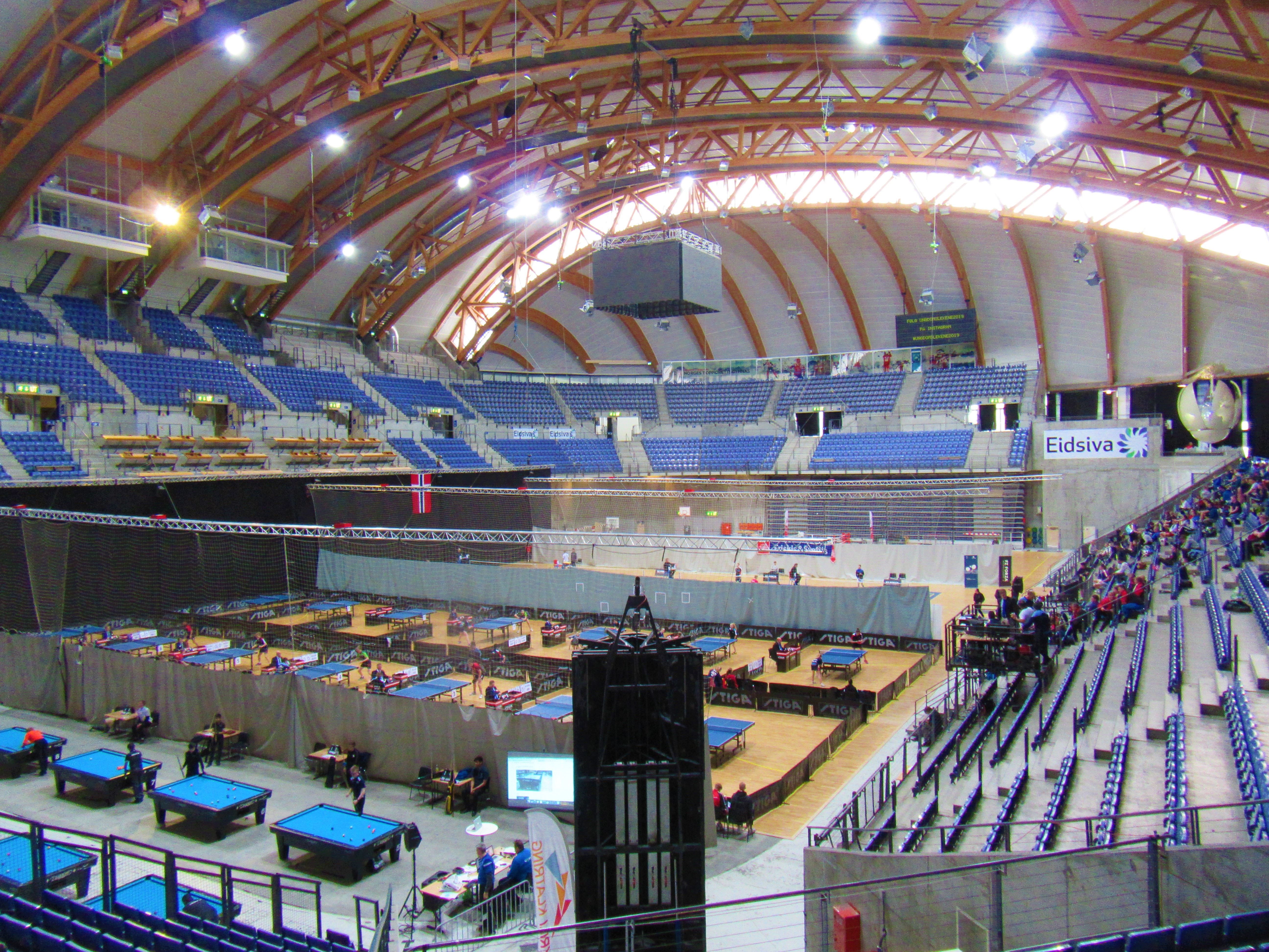 Stor innendørs arena med buet tretak, tribuner og mange bordtennisbord på banen med noen tilskuere..