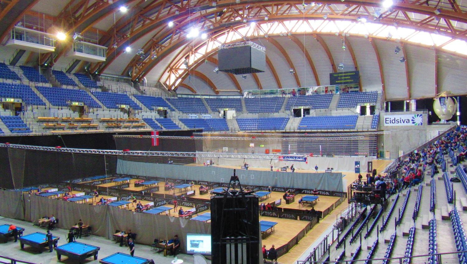 Stor innendørs arena med buet tretak, tribuner og mange bordtennisbord på banen med noen tilskuere..