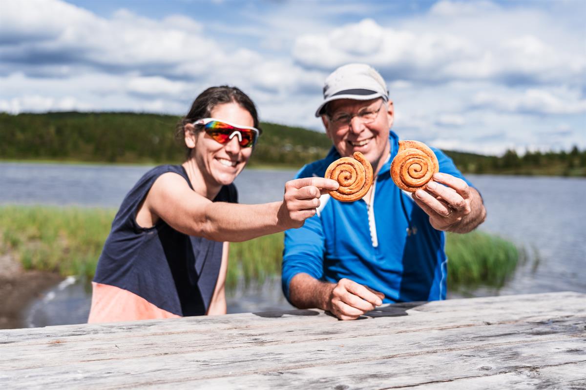 Eine Frau und ein Mann lächeln und halten zwei Zimtschnecken in die Kamera, im Hintergrund ein See.