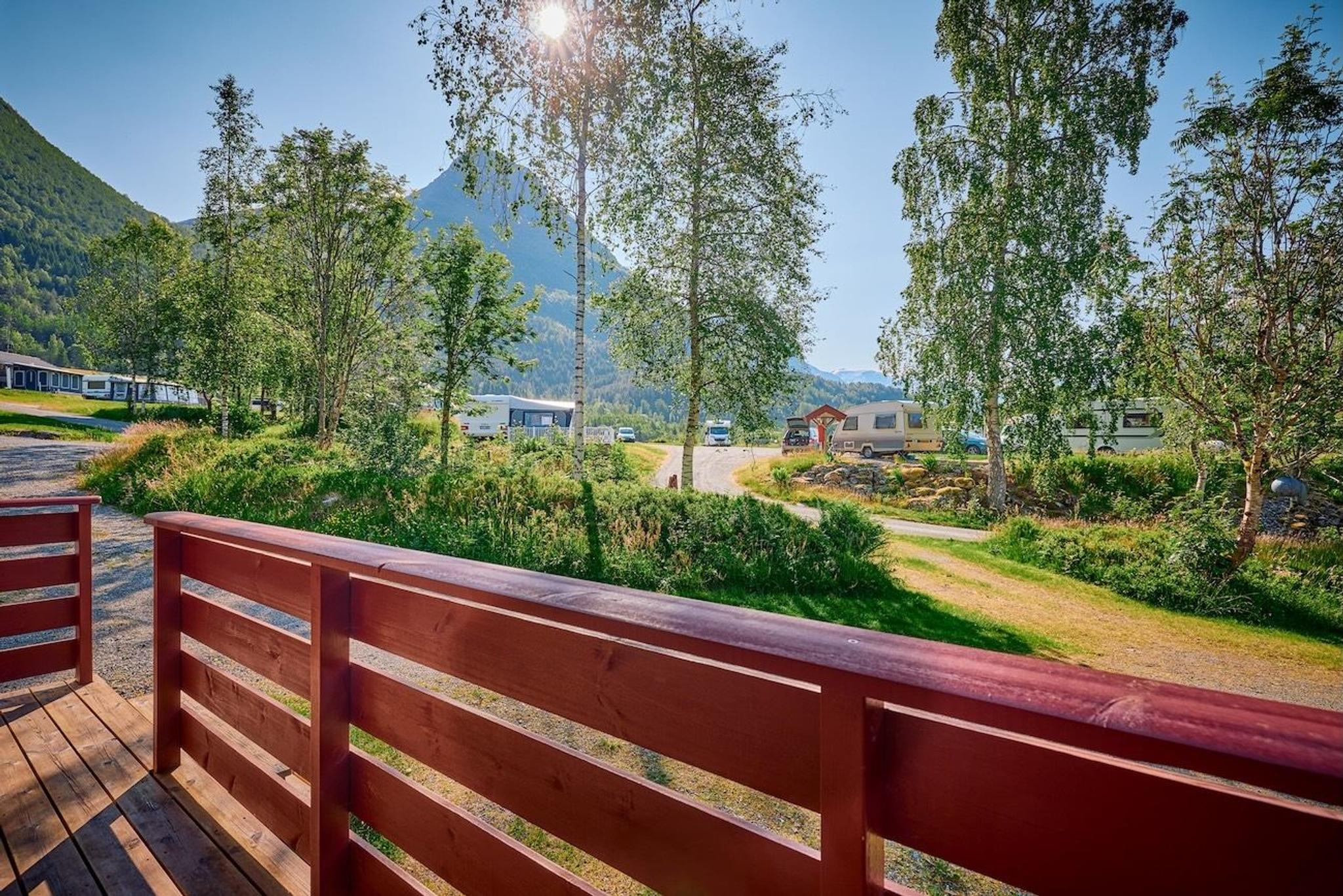 Utsikt frå treterrasse med raudt rekkverk mot campingplass med bubilar, bjørketrær og fjell i solskin..