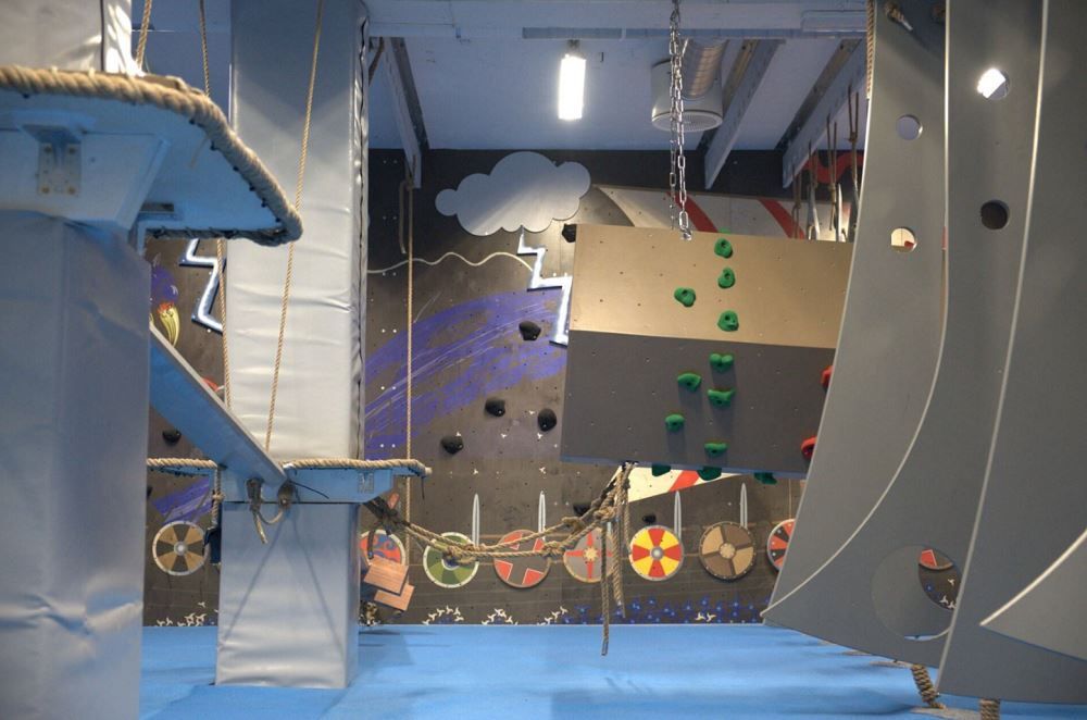 Indoor obstacle course with padded platforms, ropes, and a small climbing wall with green holds in a gym..