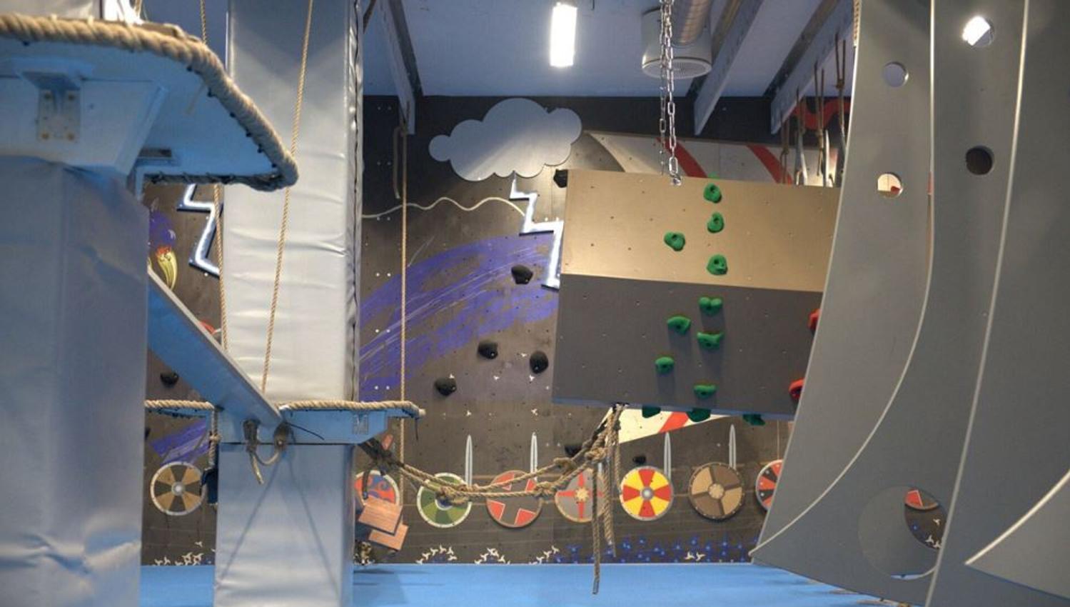 Indoor obstacle course with padded platforms, ropes, and a small climbing wall with green holds in a gym..