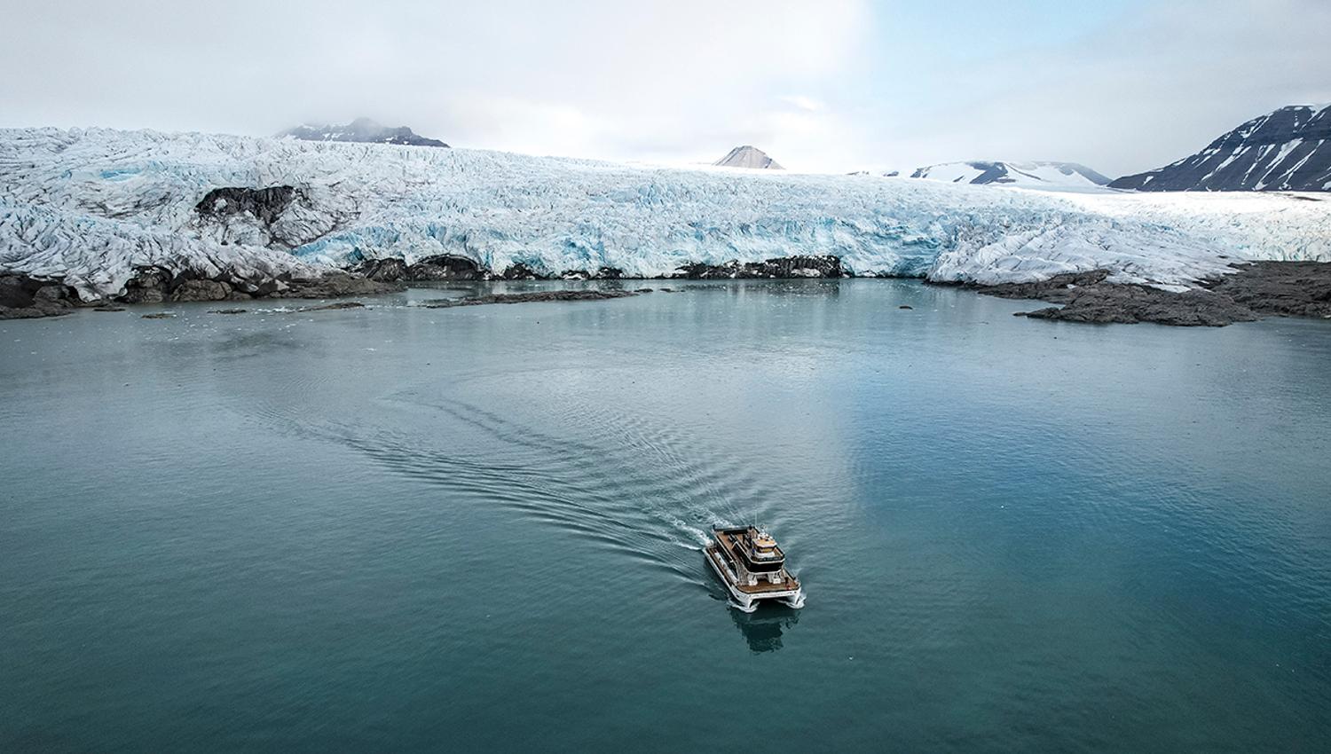 A boat sailing on a fjord in the foreground with a glacier front and mountains surrounding a bay in the background