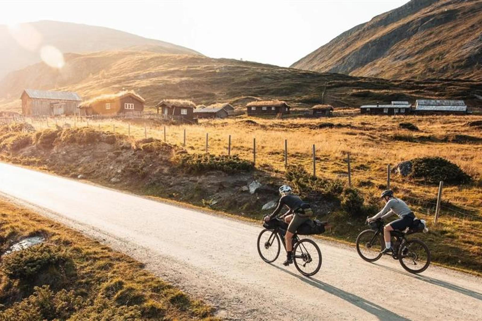 Two people cycling along a country road in the mountains, with small turf-roofed cabins in the background.