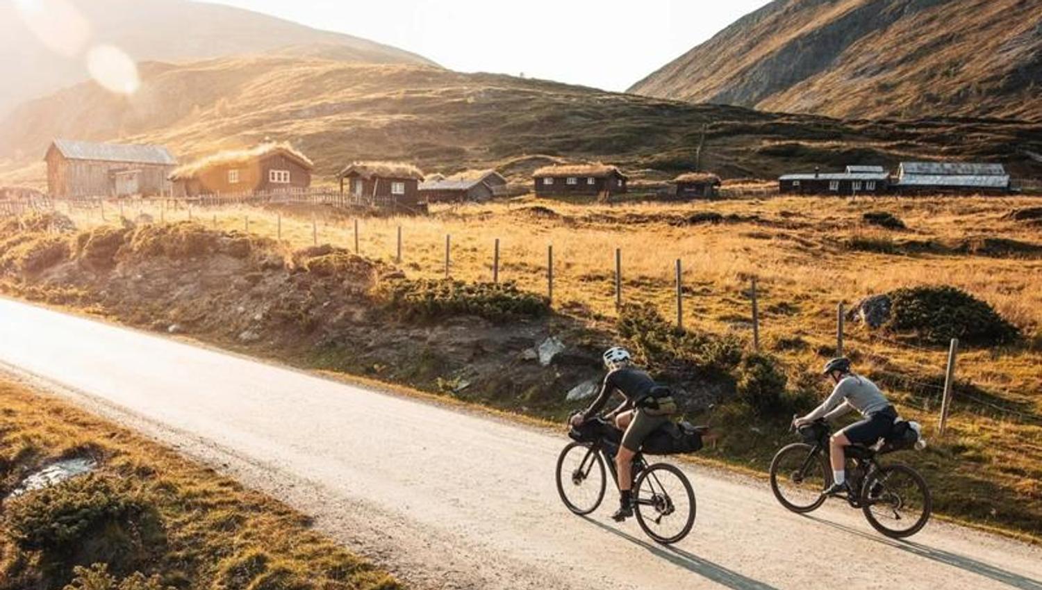 Two people cycling along a country road in the mountains, with small turf-roofed cabins in the background.