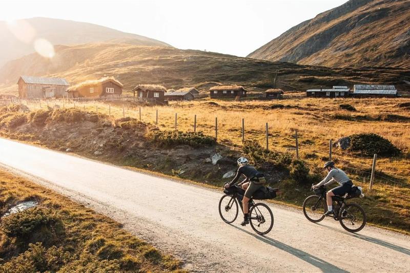 Two people cycling along a country road in the mountains, with small turf-roofed cabins in the background.