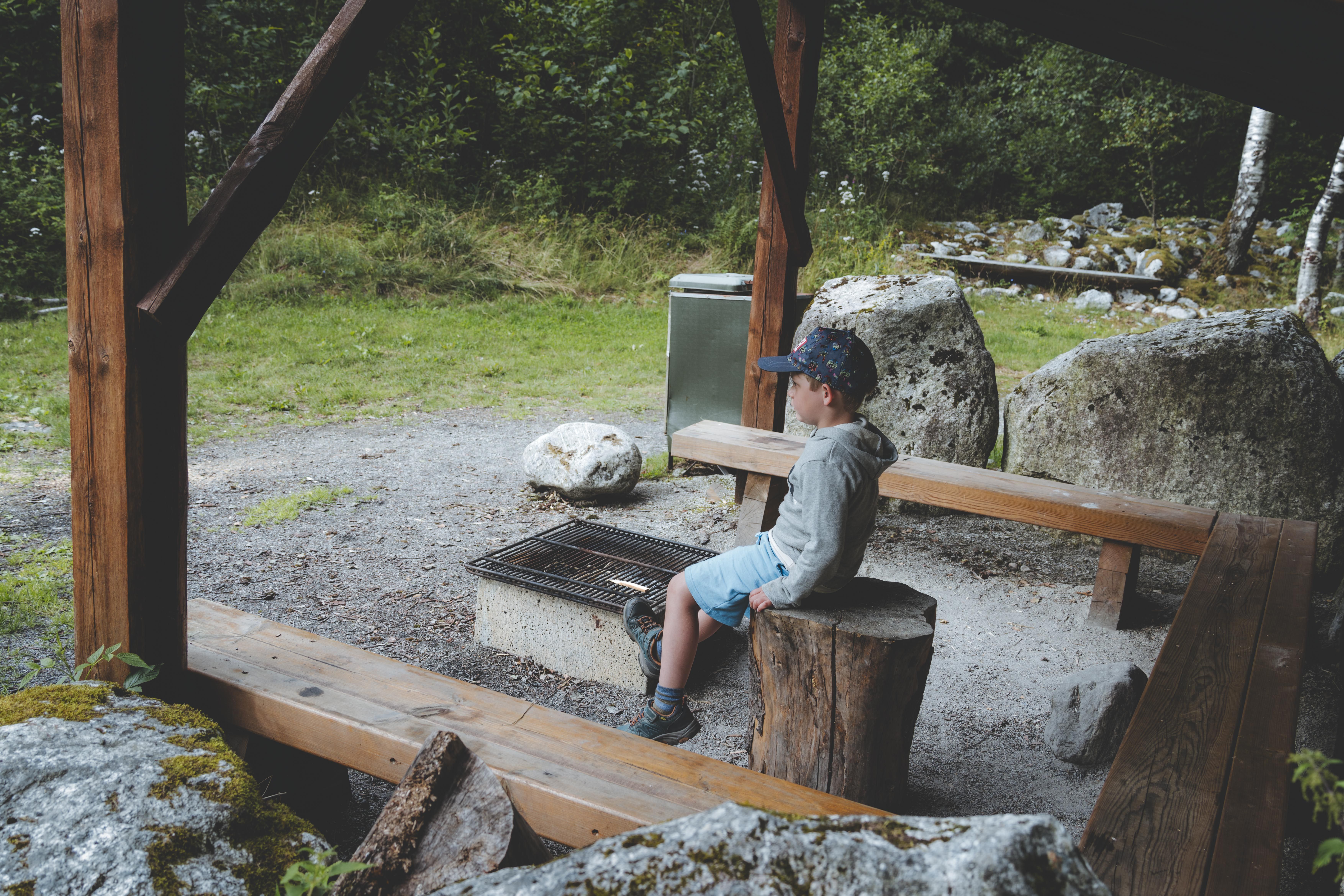 A little boy sitting on a bench in a fishing shelter, in front of a bbq area.