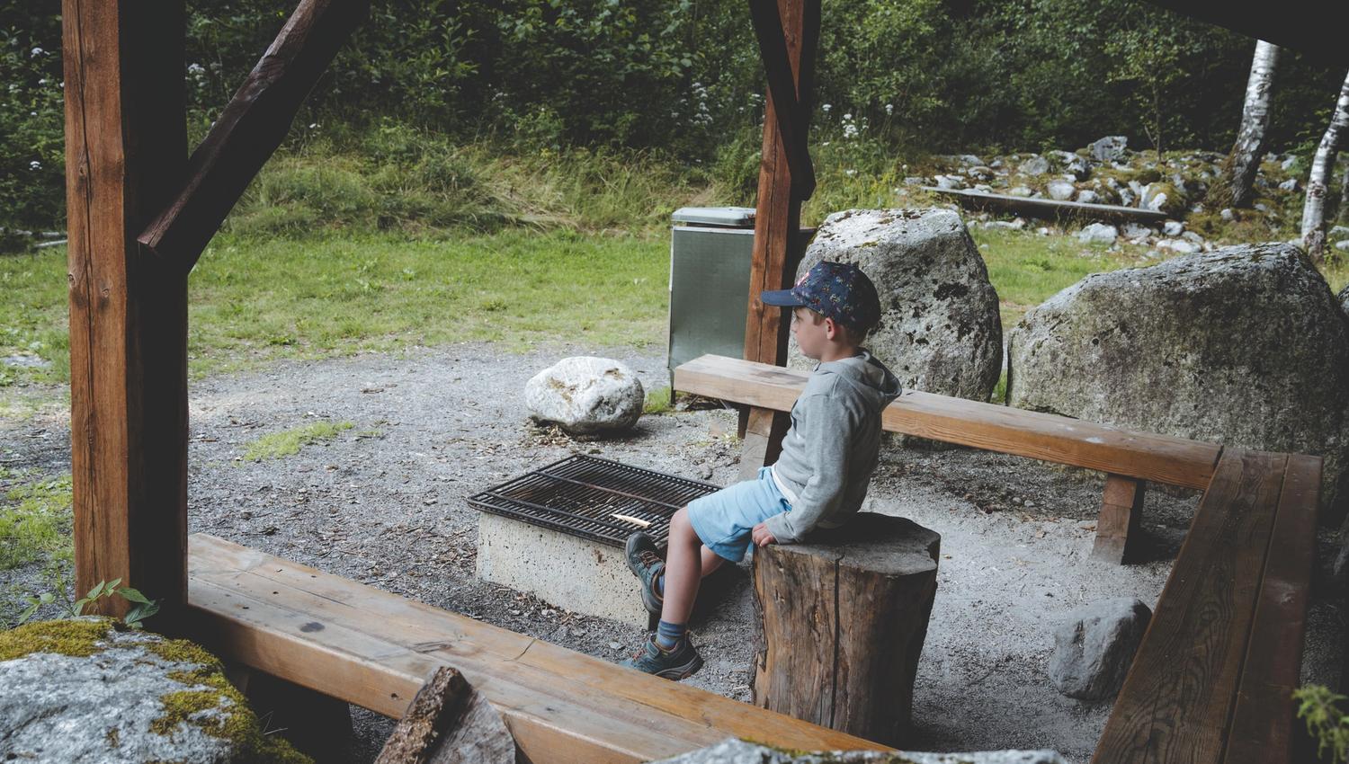 A little boy sitting on a bench in a fishing shelter, in front of a bbq area.