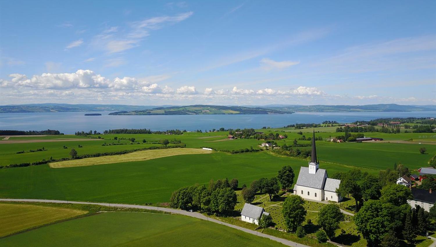 Pilegrimsleden Stange Vestbygd. Utsikt over et grønt jordbrukslandskap med åkrer, gårder og Mjøsa i bakgrunnen.