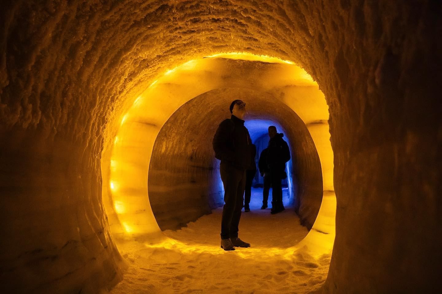 To personar står i ein rund istunnel med gul belysning, medan blått lys lyser frå opninga lenger inne..