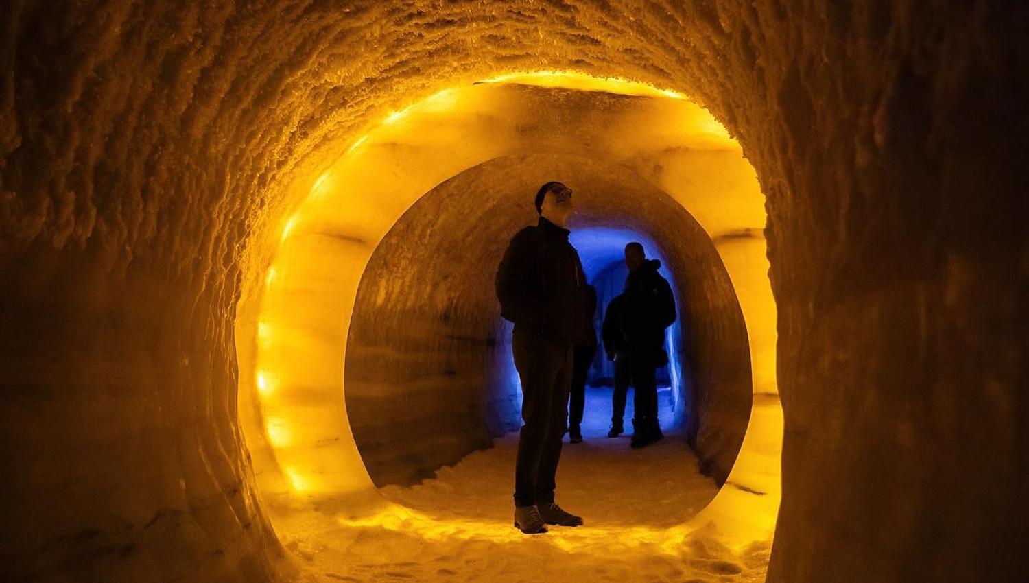 To personar står i ein rund istunnel med gul belysning, medan blått lys lyser frå opninga lenger inne..