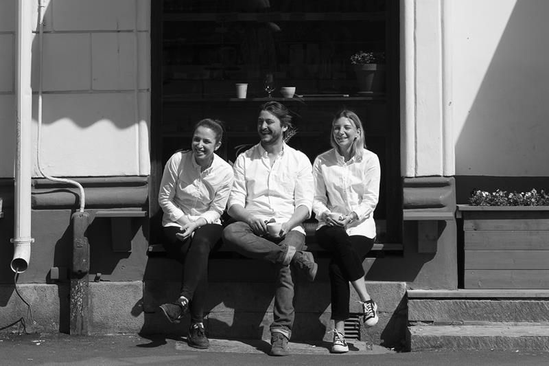 Sort-hvitt bilde. 3 smilende personer sitter utenfor cafélokale.