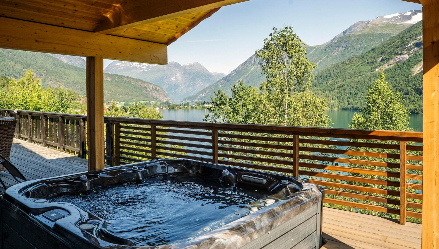 A bubbling hot tub on a wooden covered deck overlooks a calm lake, green trees, and distant mountains..