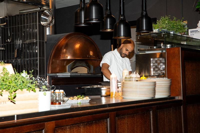 En kokk forbereder mat ved kjøkkenbenken foran en vedfyrt ovn i et moderne restaurantkjøkken.