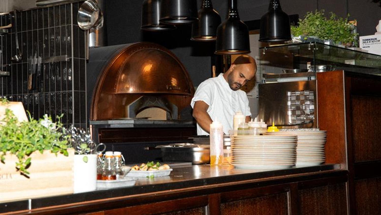 En kokk forbereder mat ved kjøkkenbenken foran en vedfyrt ovn i et moderne restaurantkjøkken.