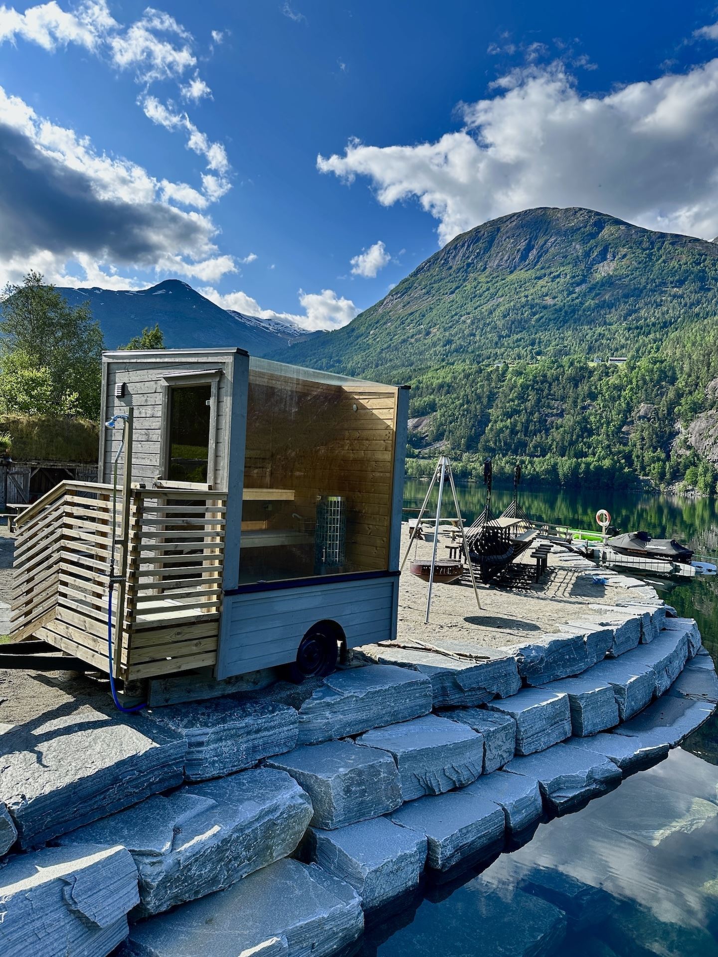 Small wooden sauna cabin on a stone dock beside a calm lake, with forested mountains and clouds overhead..