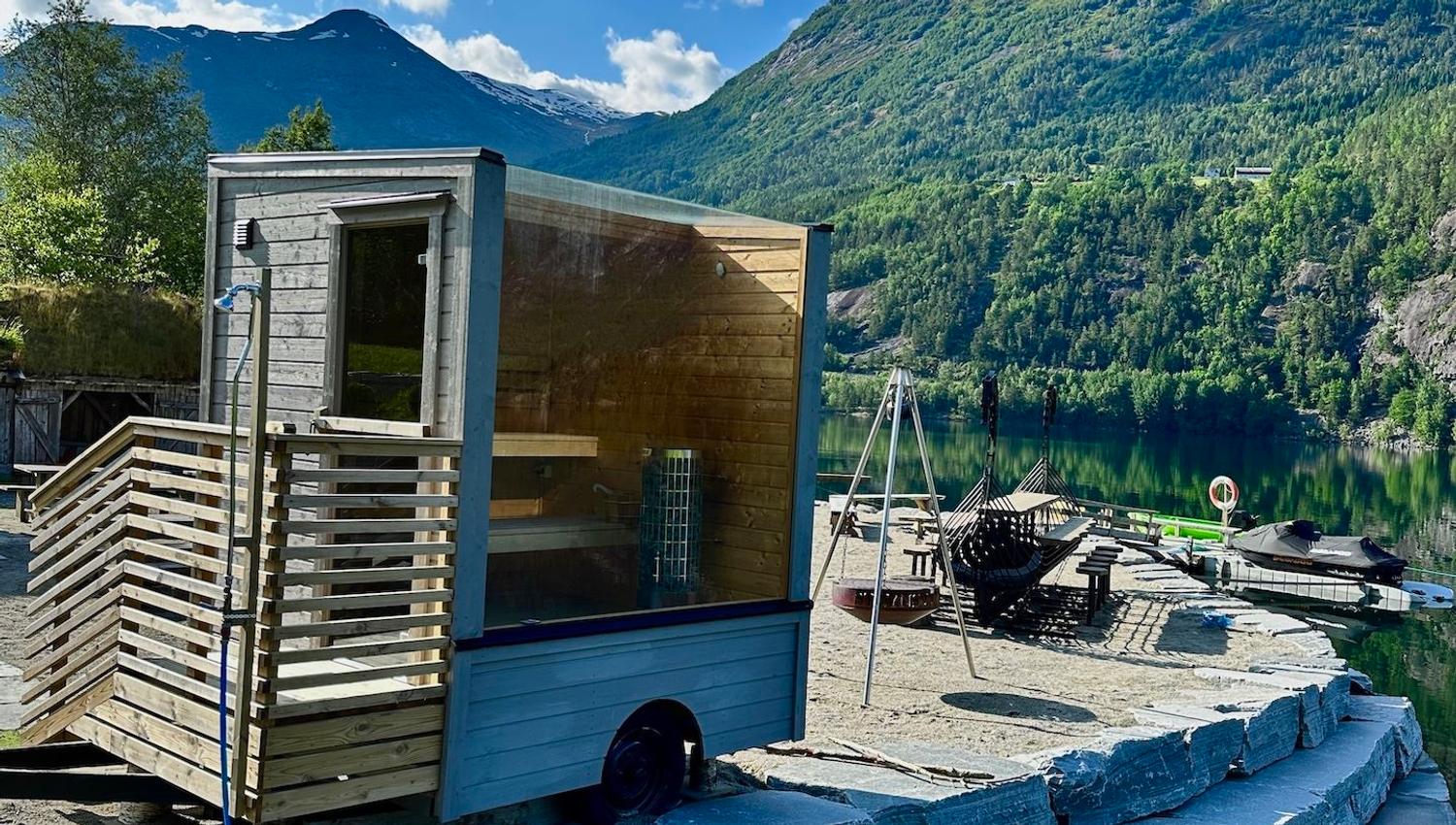 Small wooden sauna cabin on a stone dock beside a calm lake, with forested mountains and clouds overhead..