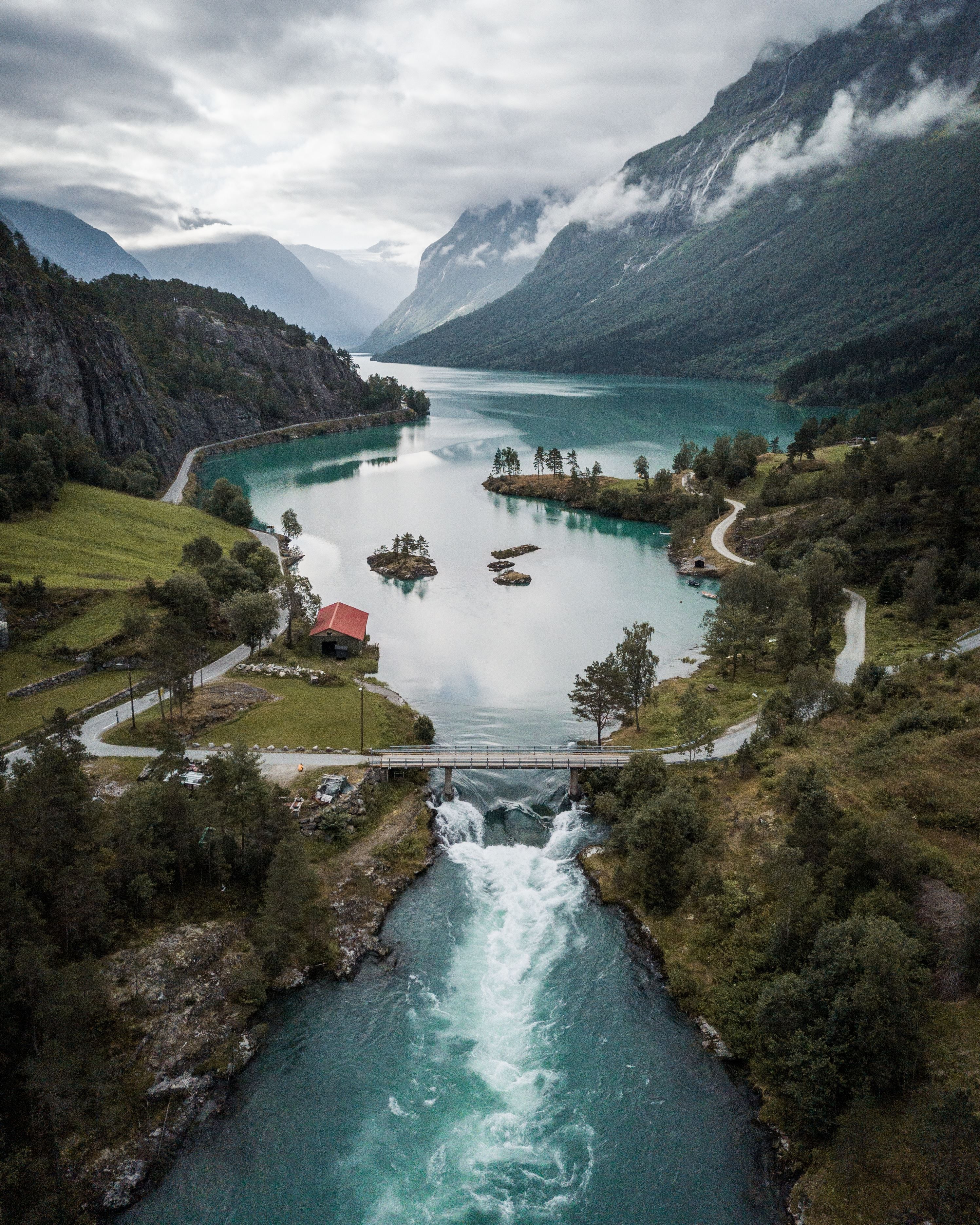 Aerial view of turquoise Lovatnet lake between mountains, with a river, small bridge, and a red-roof cabin below..