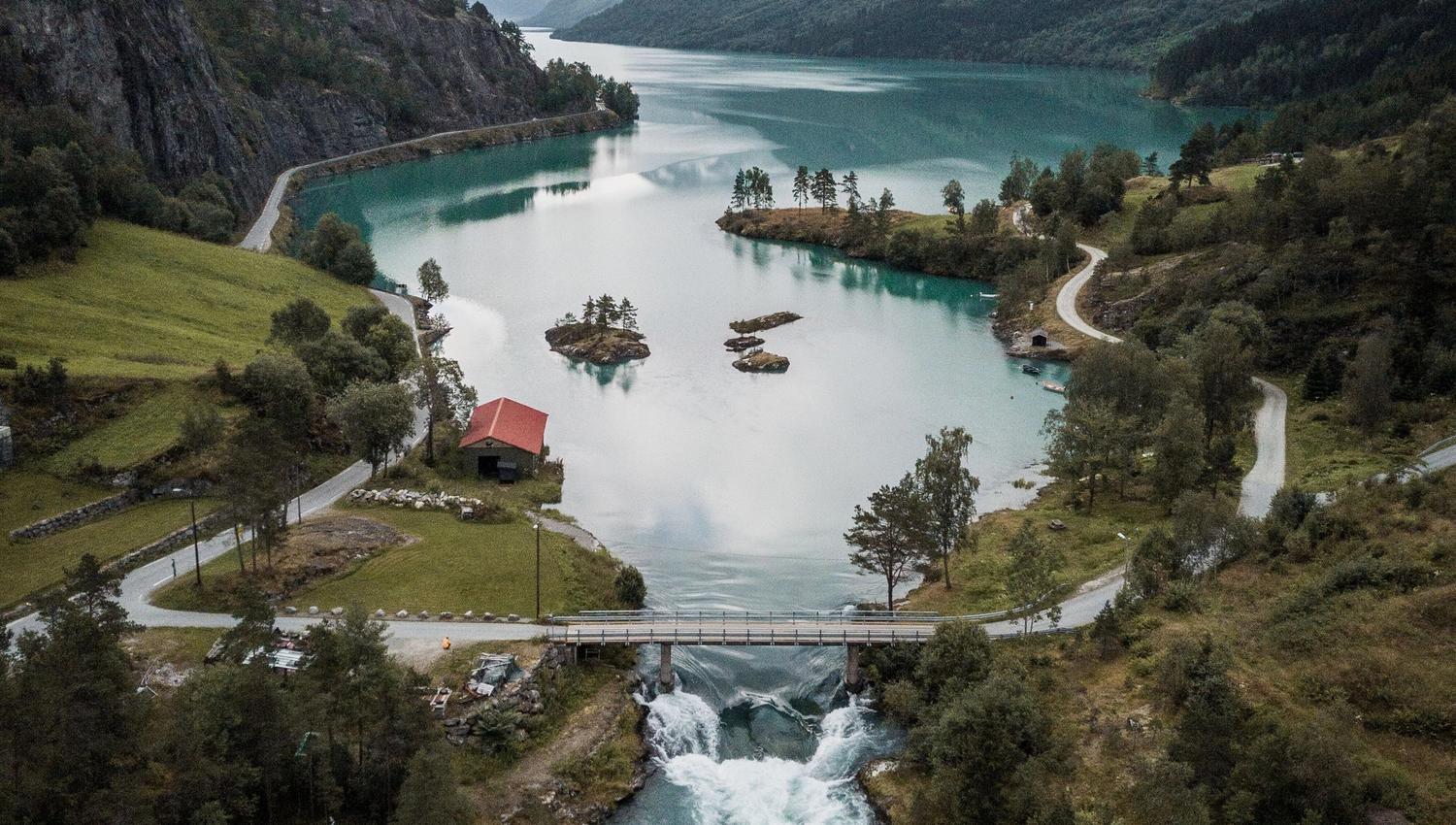 Aerial view of turquoise Lovatnet lake between mountains, with a river, small bridge, and a red-roof cabin below..