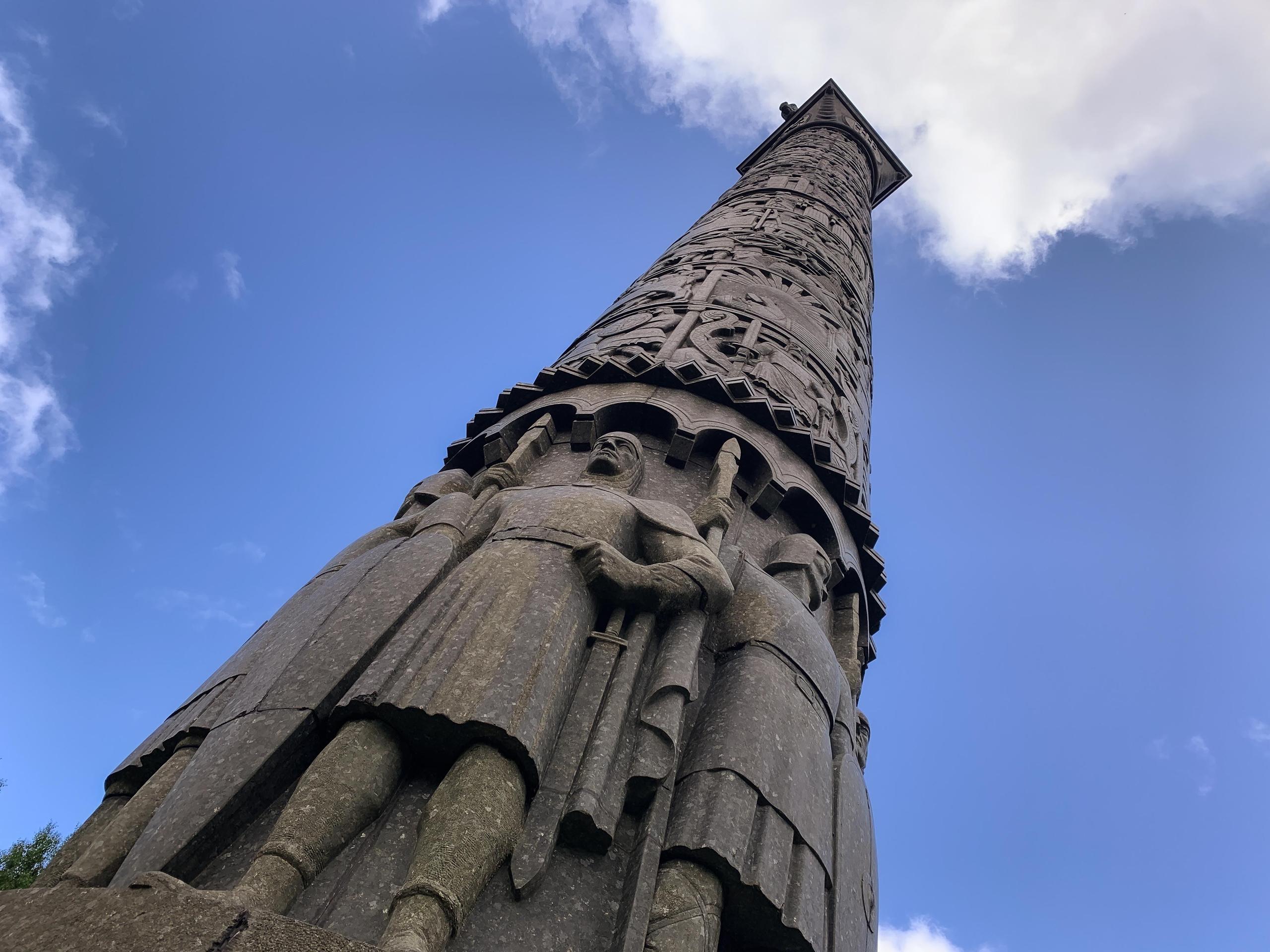 Low-angle view of a tall carved stone monument with robed statues at the base against a blue, cloudy sky..