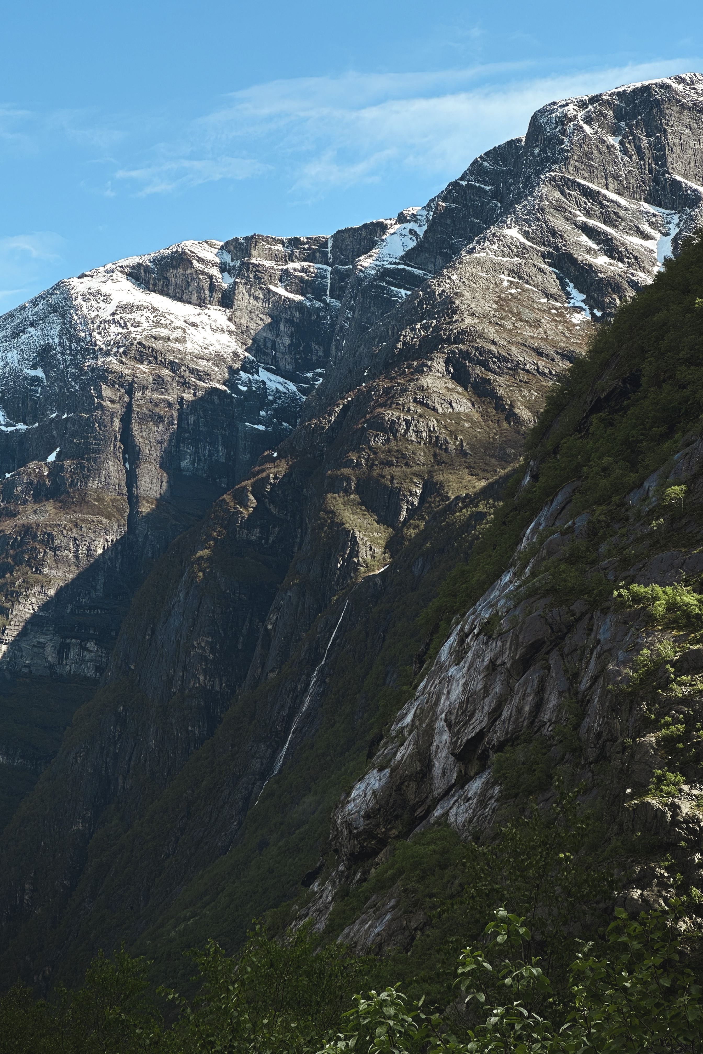 Steep rocky mountains with patches of snow rise above a green forested slope under a clear blue sky..