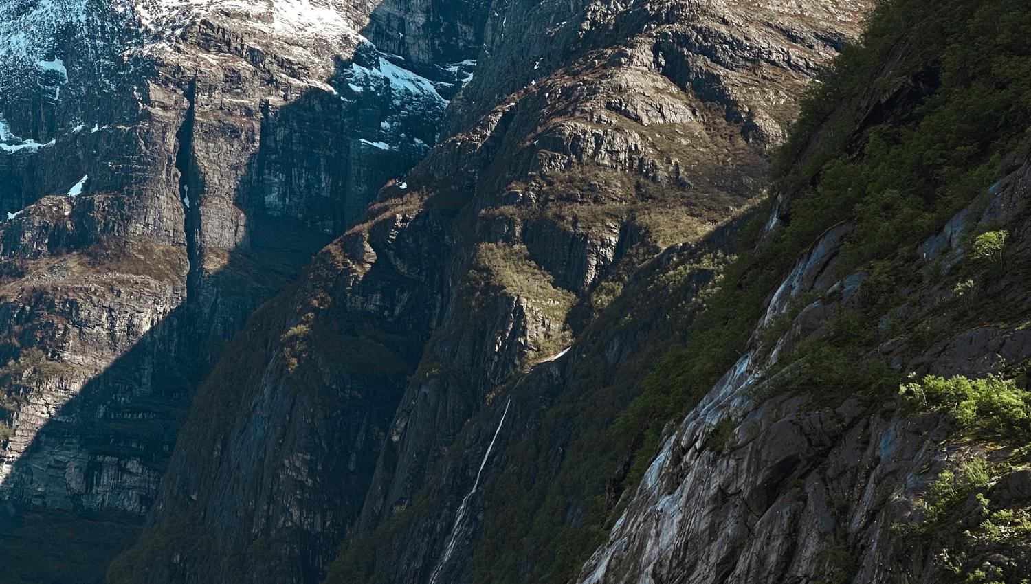 Steep rocky mountains with patches of snow rise above a green forested slope under a clear blue sky..