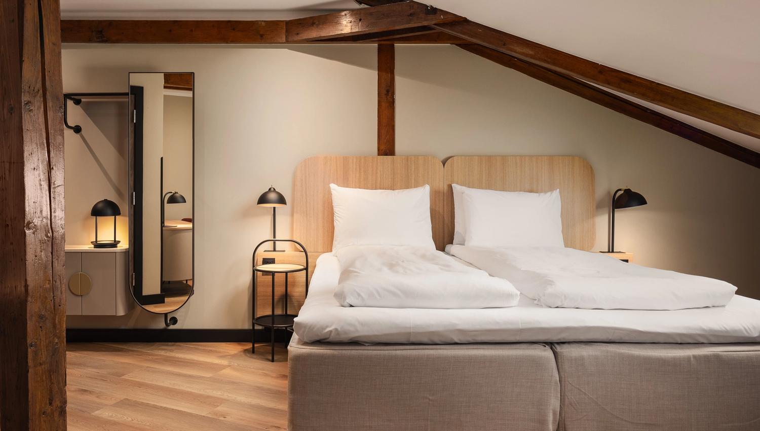 A hotel room interior with a double bed, a mirror, and a brown ceiling beam running along the ceiling.