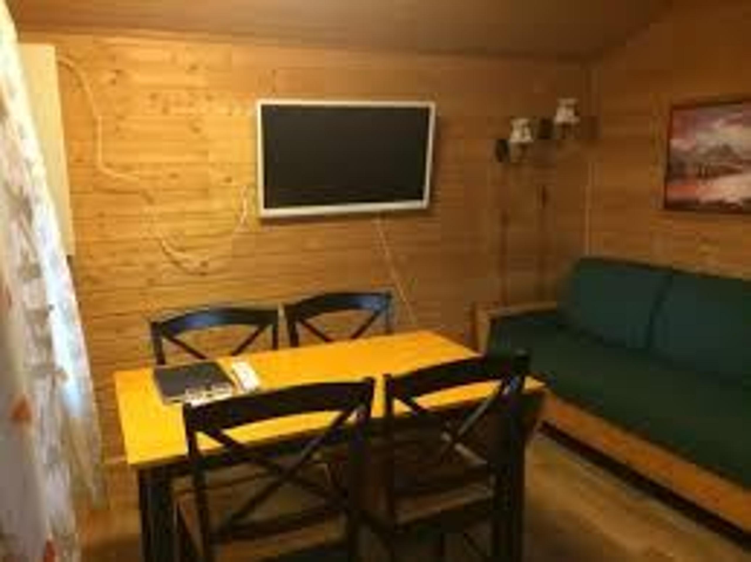 Wood-paneled cabin room with a dining table, black chairs, wall-mounted TV, and a green bench sofa.