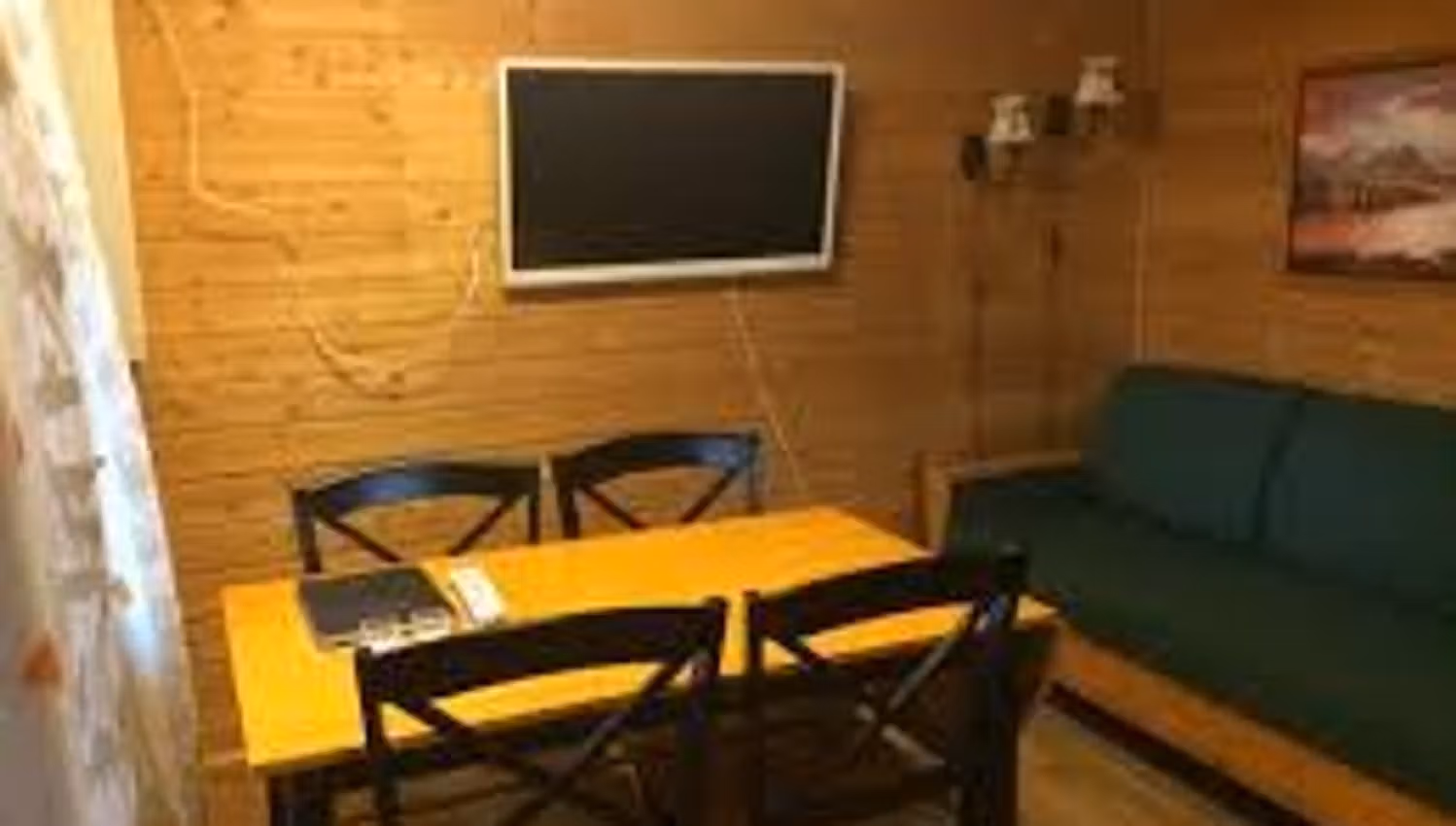 Wood-paneled cabin room with a dining table, black chairs, wall-mounted TV, and a green bench sofa.