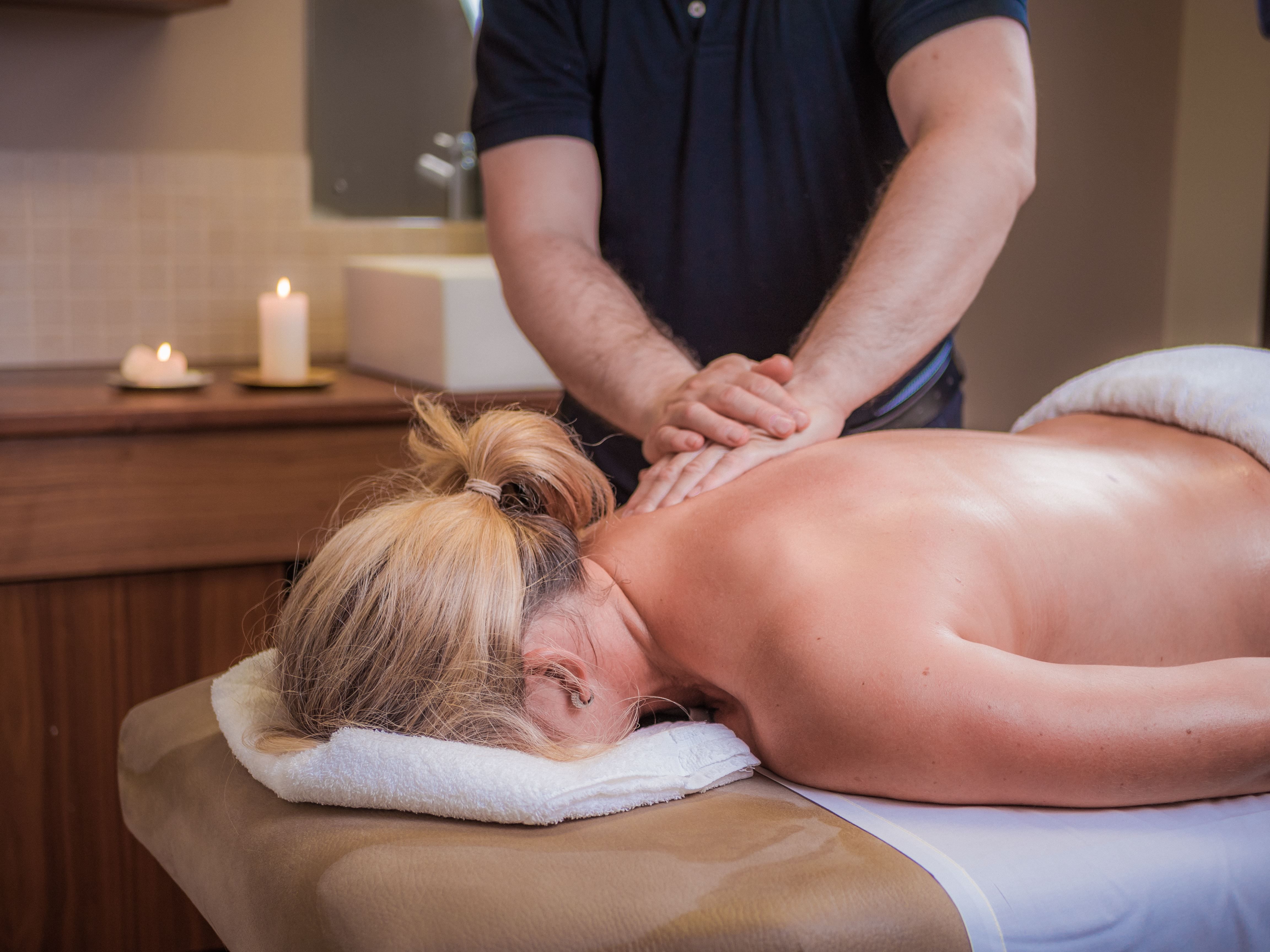 Massage therapist presses hands on a client's bare back on a spa table, with towels and candles in the room.