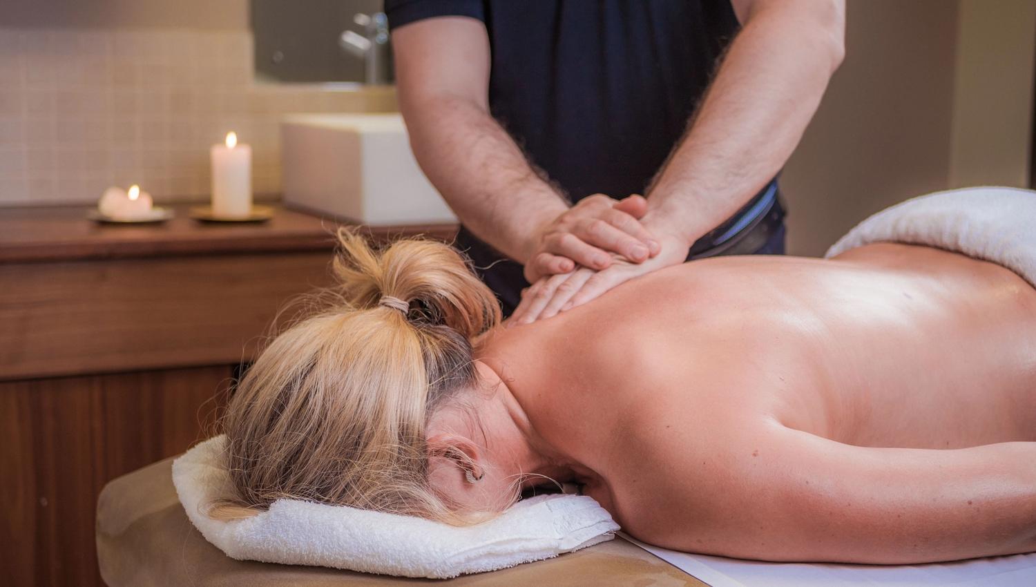 Massage therapist presses hands on a client's bare back on a spa table, with towels and candles in the room.