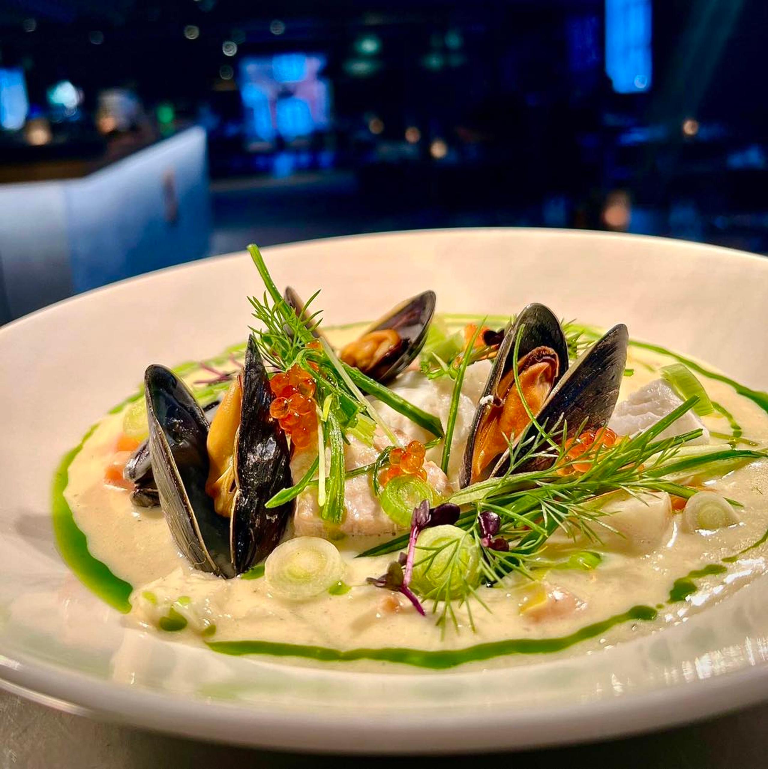 Kremet suppe med blåskjell og friske urter på en hvit tallerken, med restaurantlokalet i bakgrunnen.