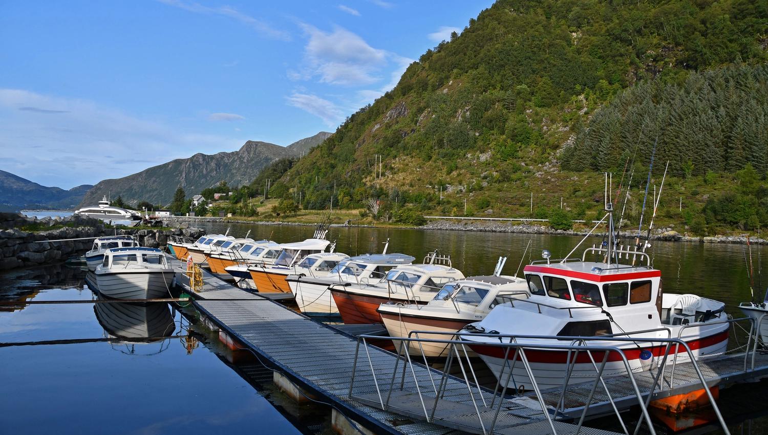 Several small motorboats moored along a floating dock on calm water, with green mountains and a blue sky behind..