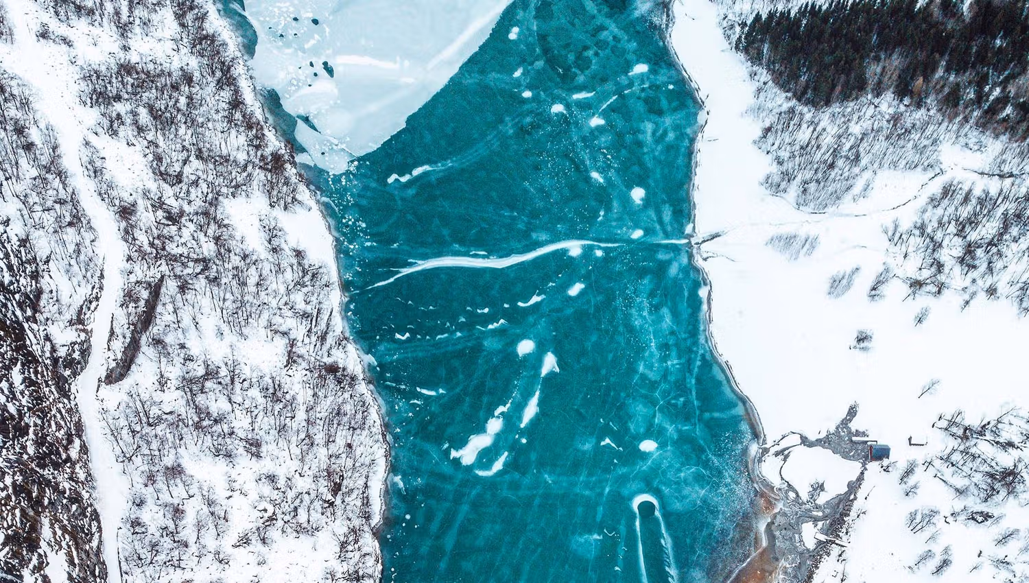 Aerial view of a turquoise frozen river with cracks, bordered by snowy forests and a small building at right..