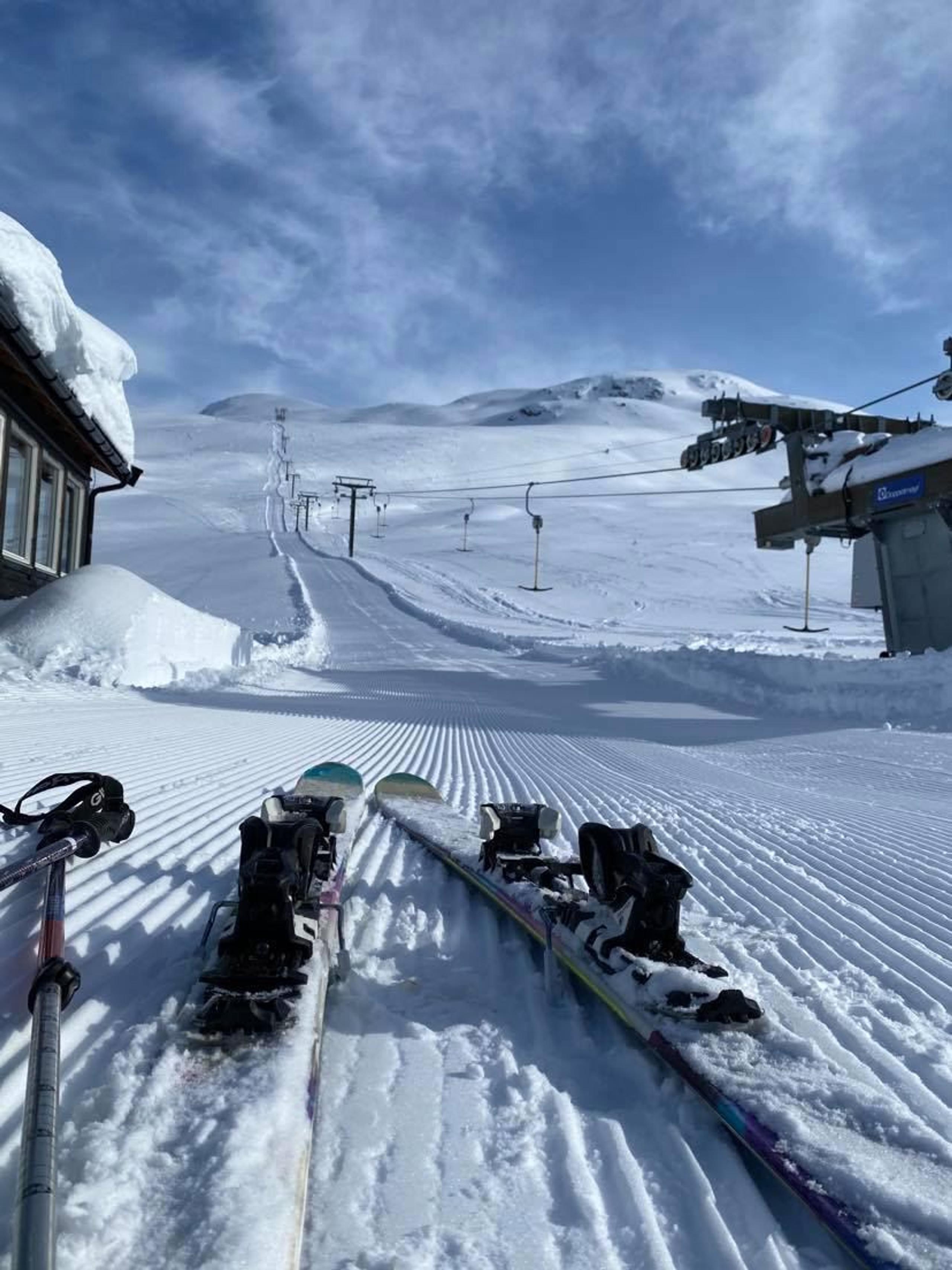 Two skis rest on a groomed snowy ski run with chairlift cables and distant mountains under a blue sky.