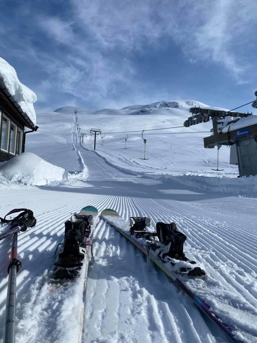 Two skis rest on a groomed snowy ski run with chairlift cables and distant mountains under a blue sky.