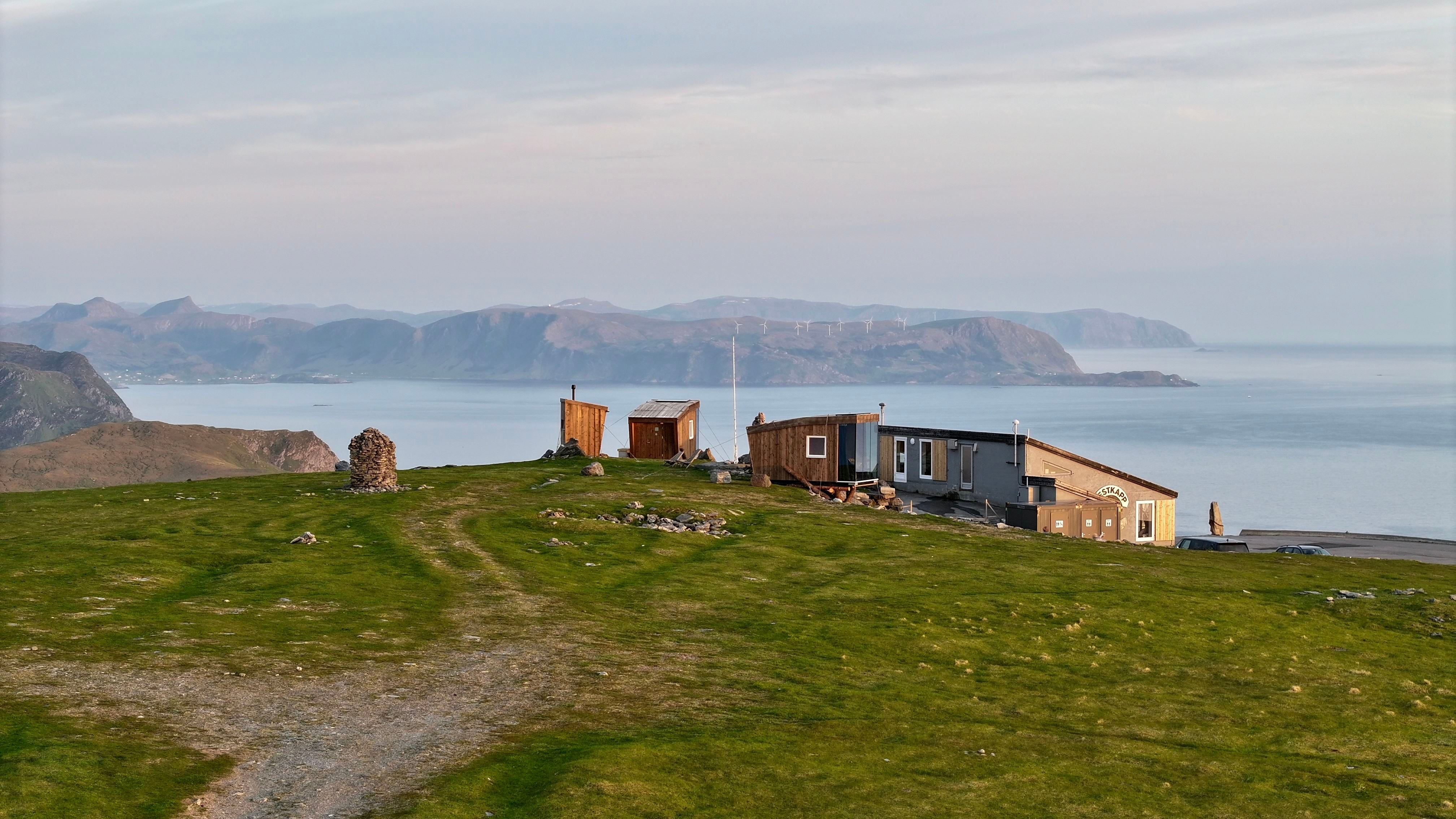 Grøn grasbakke med sti mot ei lita hytte og uthus ved fjord, med fjelløy og roleg hav i bakgrunnen..
