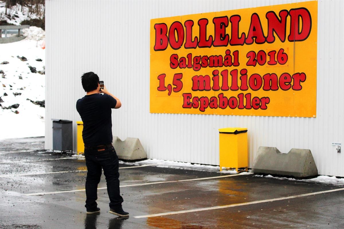 A person stands outside a building taking a photo of a large wall sign displaying the name “Bolleland” and sales information.