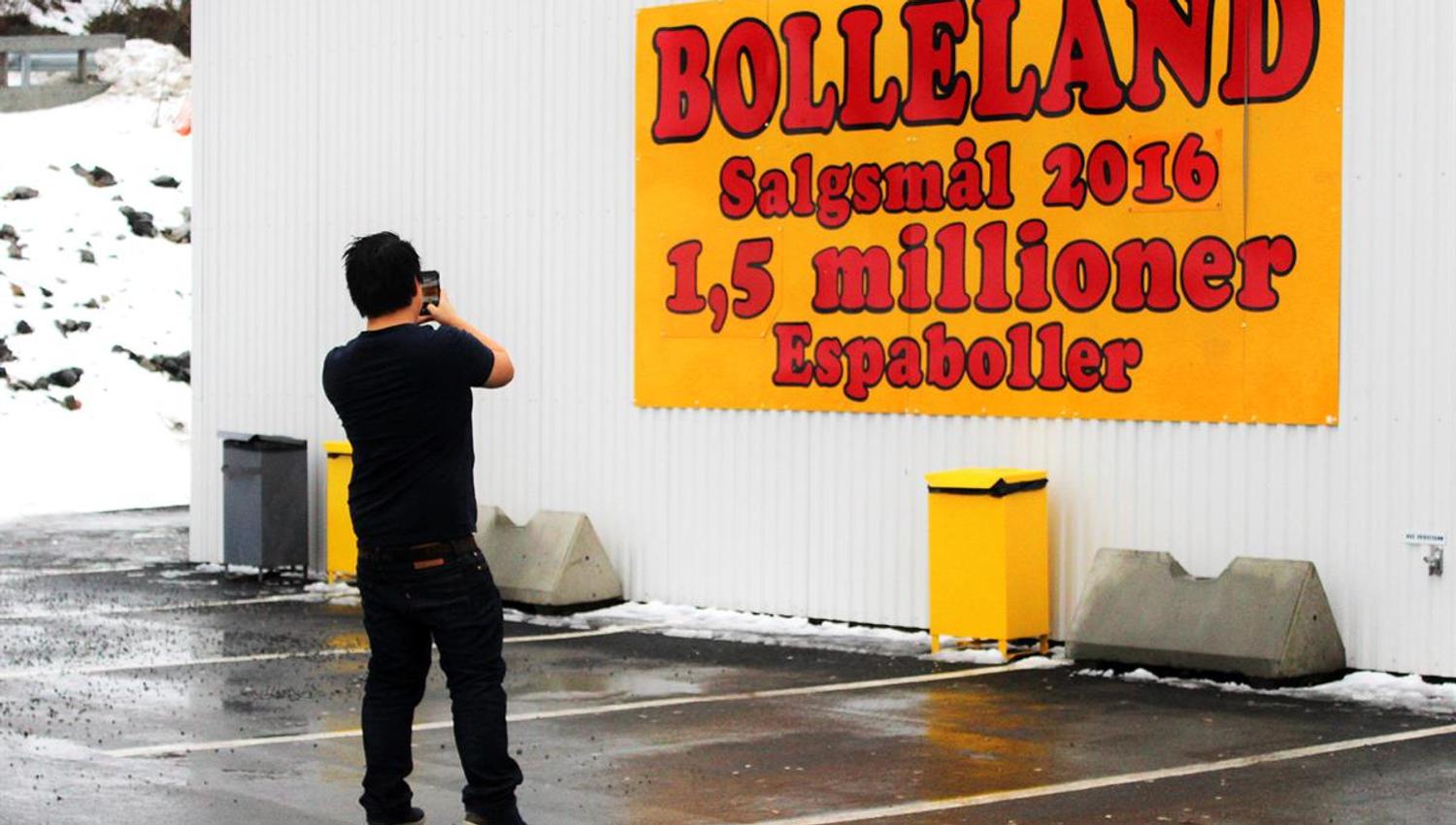 A person stands outside a building taking a photo of a large wall sign displaying the name “Bolleland” and sales information.