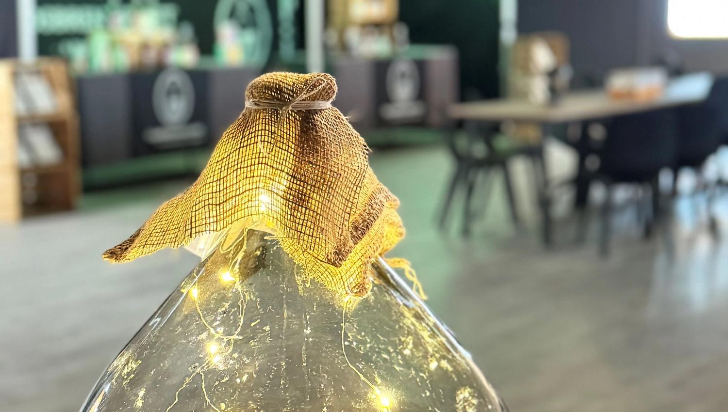 Close-up of a large glass jar filled with warm string lights, topped with burlap fabric in a blurred indoor room.