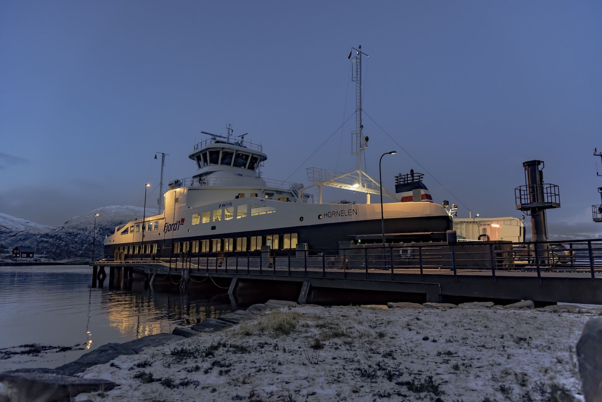 A large passenger ferry docked at a pier at dusk, with snowy shoreline in the foreground and calm water nearby..