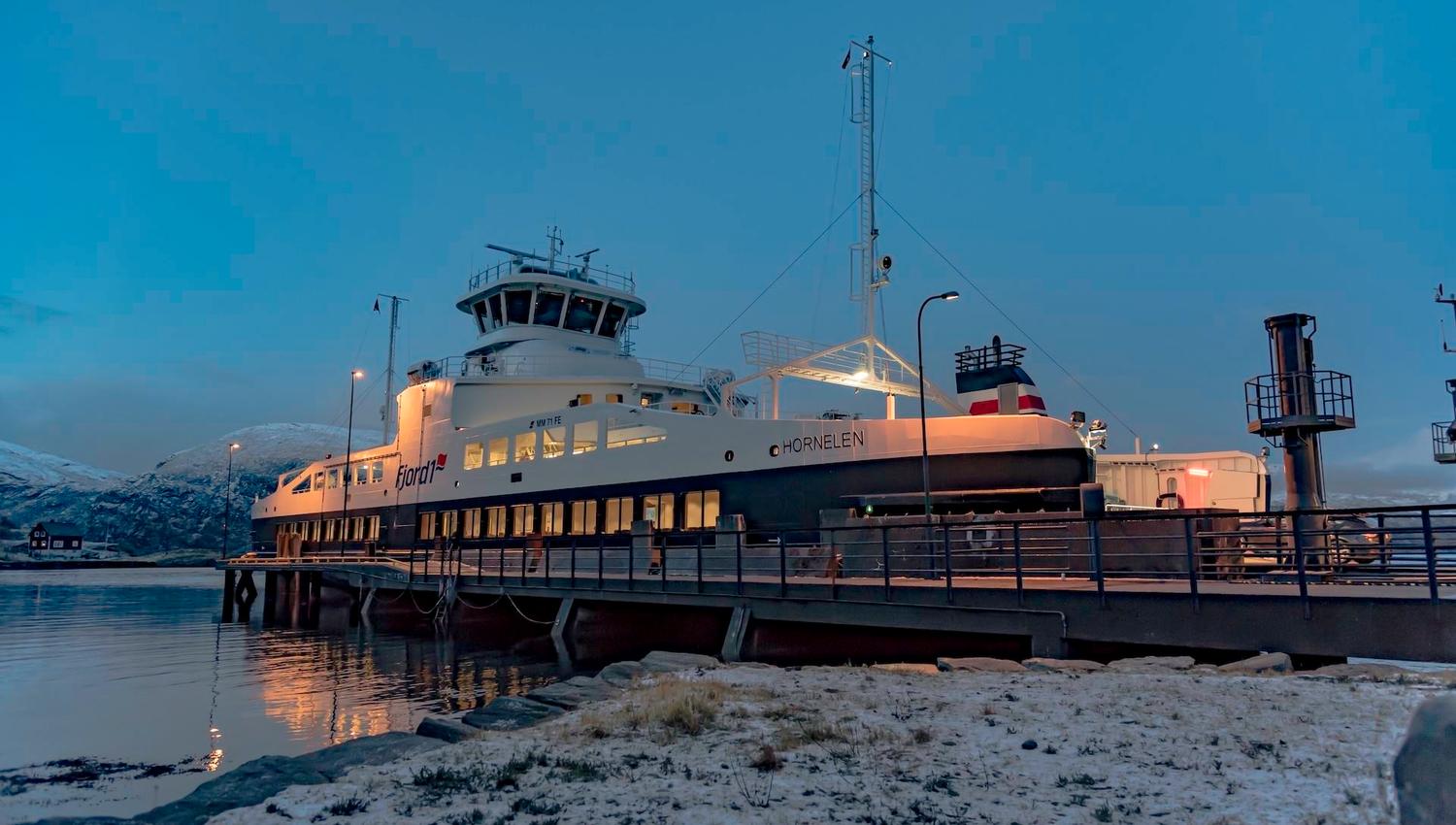 A large passenger ferry docked at a pier at dusk, with snowy shoreline in the foreground and calm water nearby..