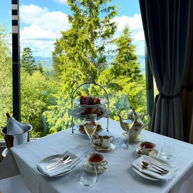 Et dekket bord for afternoon tea med utsikt over grønn skog og åser gjennom store vinduer.