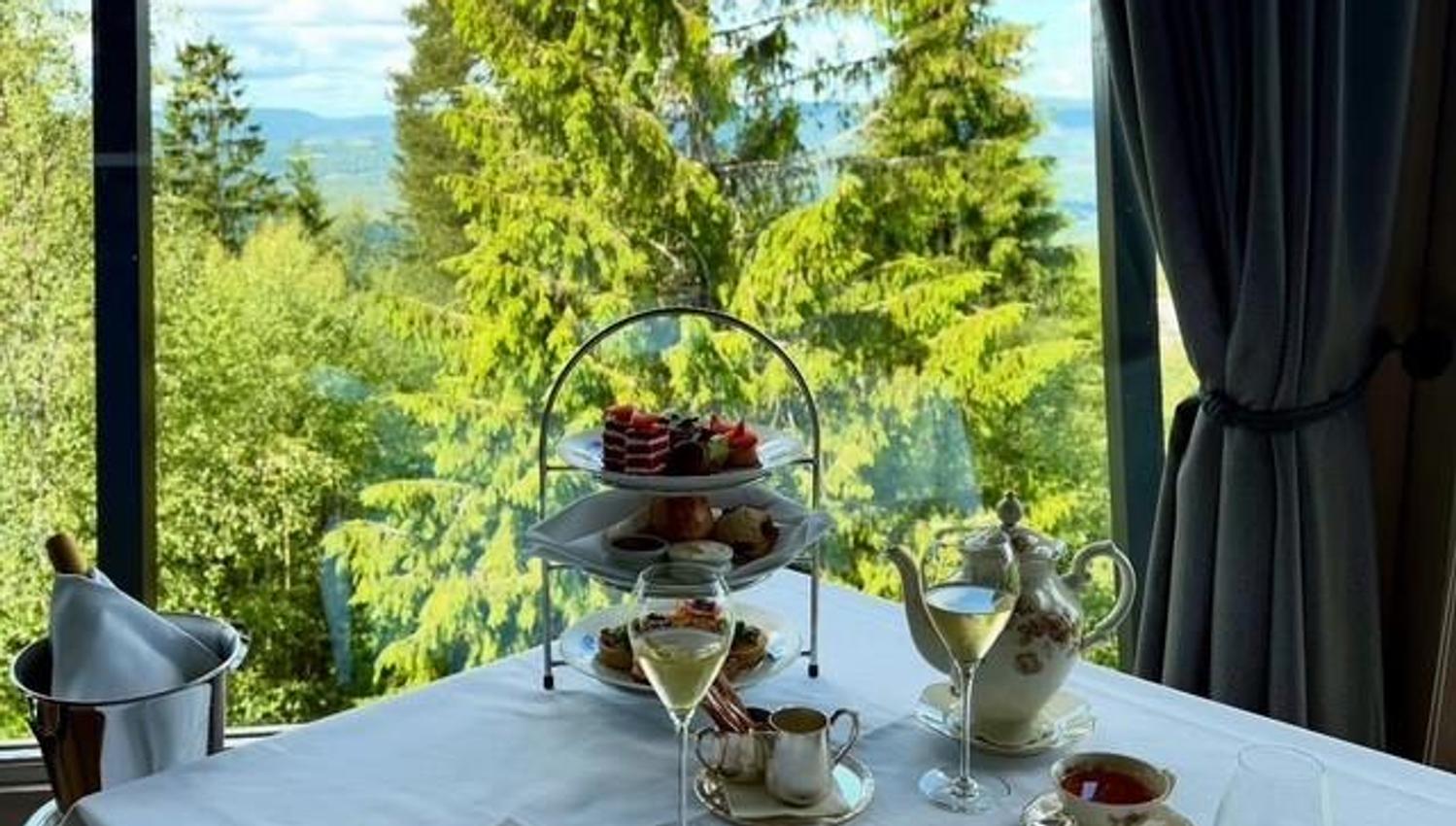 Et dekket bord for afternoon tea med utsikt over grønn skog og åser gjennom store vinduer.