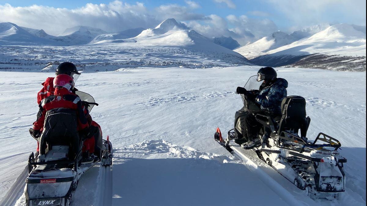 Gjester sitter på snøskutere og nyter utsikten over fjellene